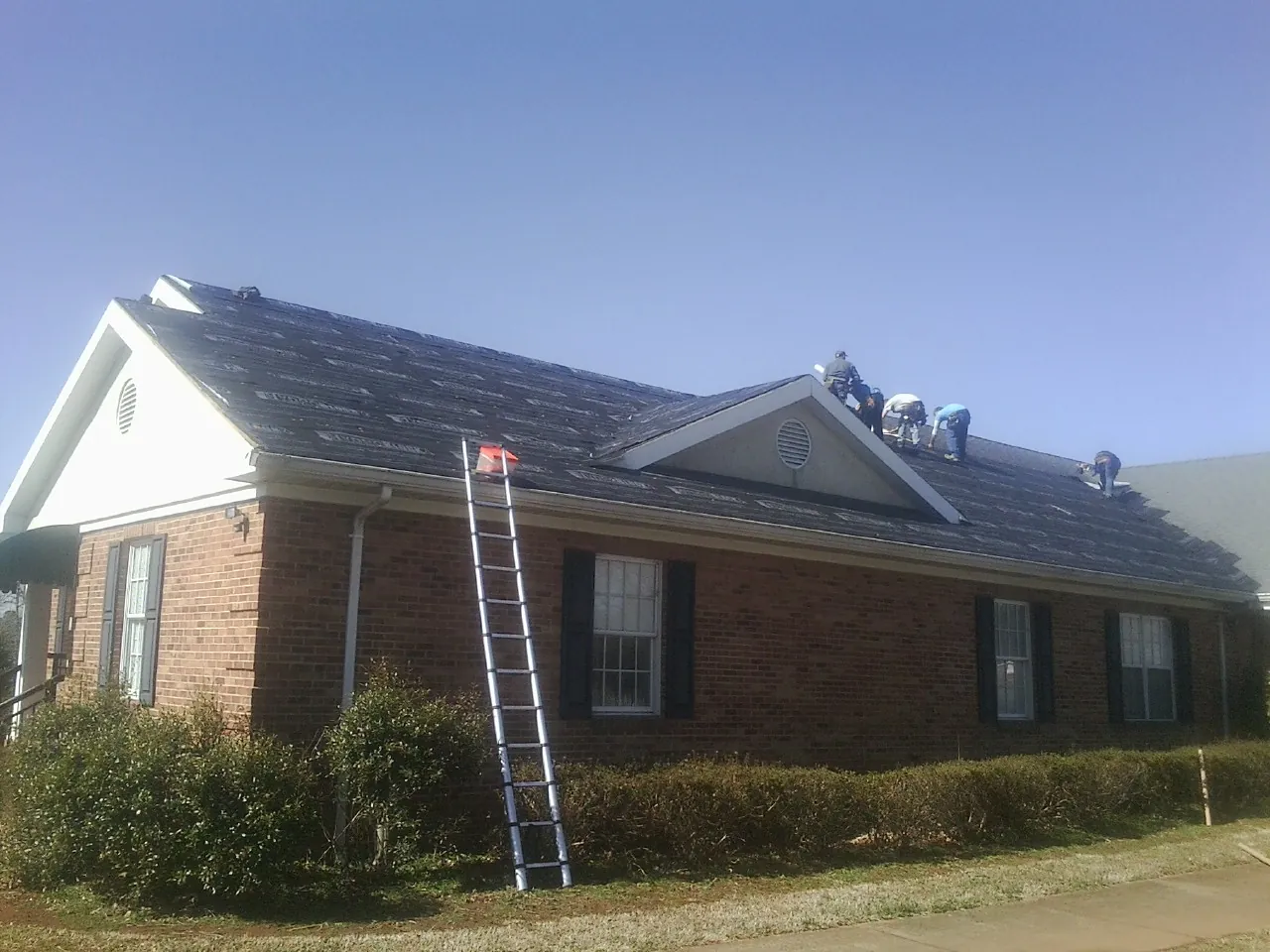 Crew tearing shingles off a brick ranch roof with a ladder set — Emergency Roof Repair work by our crew in Little Orleans