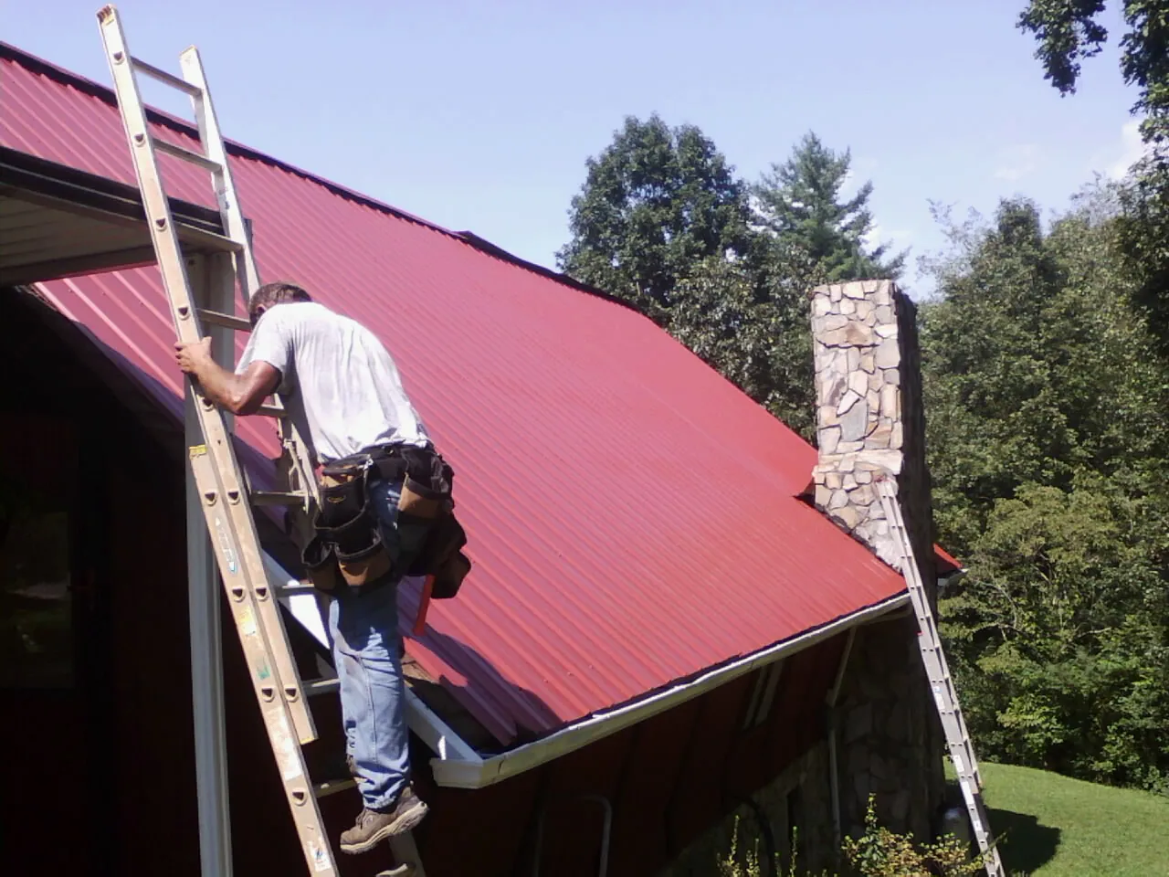 Roofer on a ladder beside a red metal roof and stone chimney — Soffit & Fascia Repair by Roofing Brothers in Little Orleans